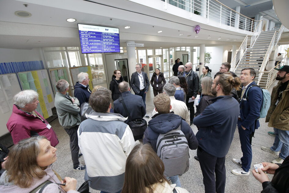 Gruppe von Menschen steht in einem hellen Foyer vor einem Bildschirm und hört einer sprechenden Person zu.