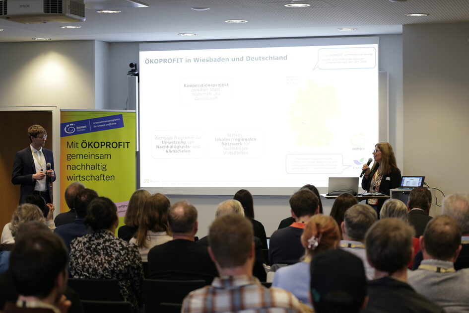 Publikum von hinten schaut auf eine weiße Leinwand sowie eine Person mit Mikrofon und ein gelbes Banner.