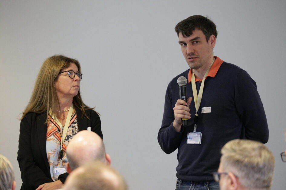 Zwei Personen mit Namensschildern stehen vor einer Gruppe, eine Person hält ein Mikrofon.