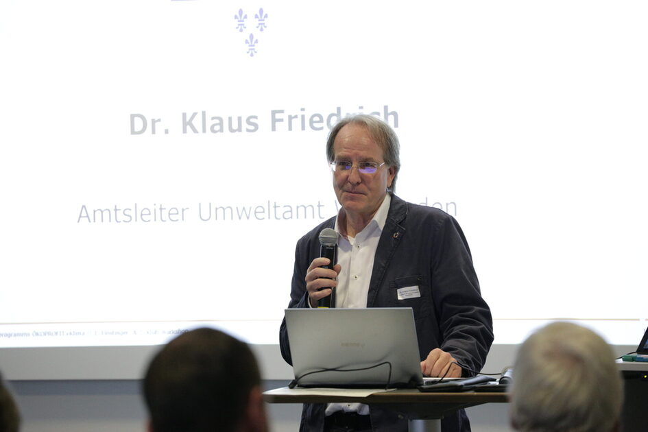 Mann mit grauen Haaren und Anzugjacke an einem Stehpult, in der Hand hält er ein Mikrofon.