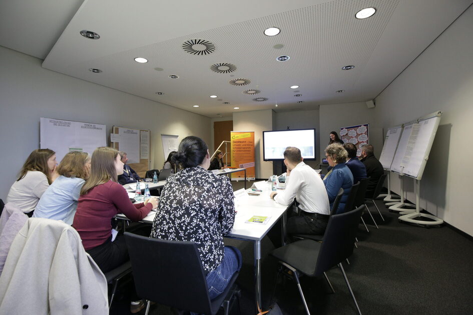 Menschen von hinten zu sehen, sitzen an Tischen zu einem U geformt und schauen auf eine Präsentation auf einen Bildschirm. An den Wänden stehen Flipcharts mit Notizen.