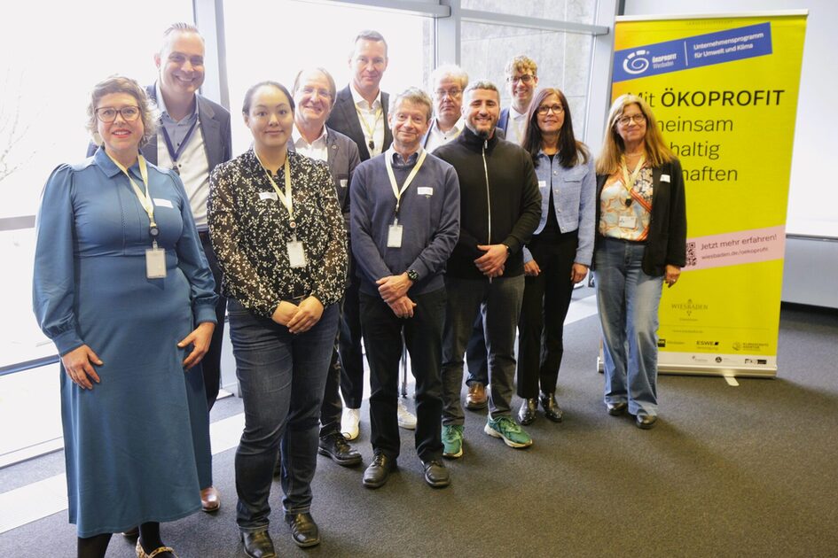 Menschengruppe mit Namenschilderm steht vor einem großen Fenster auf grauem Teppich, rechts neben der Gruppe steht ein gelbes Banner mit der Aufschrift ÖKOPROFIT.