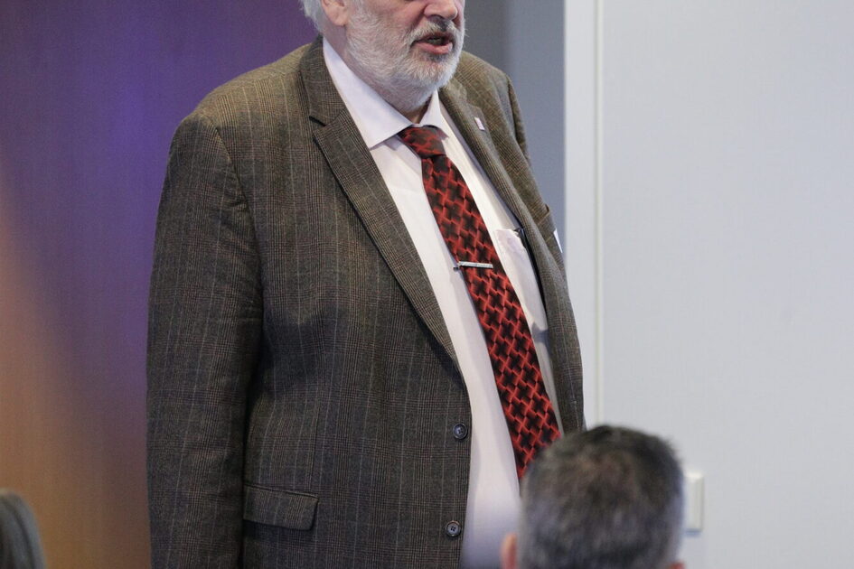 Älterer Mann mit weißem Haar, kariertem Sakko und roter Krawatte steht vor einer Gruppe sitzender Personen in einem Raum.