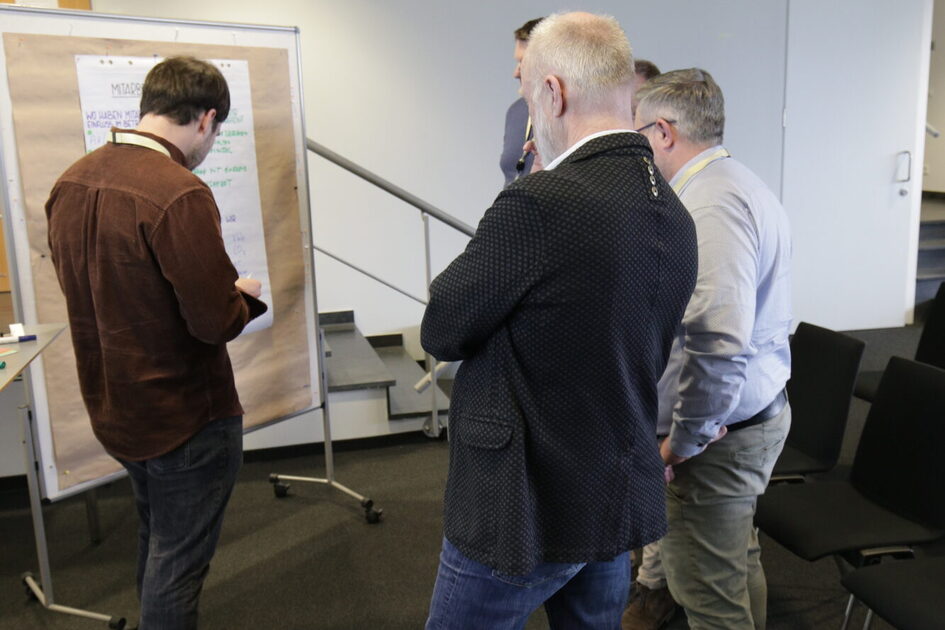 Vier Männer stehen vor einem Flipchart, einer schreibt darauf, die anderen schauen zu.