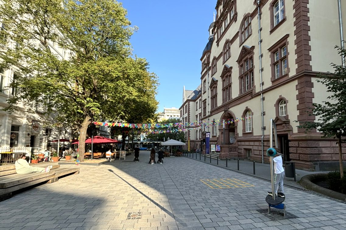 Хората се разхождат и сядат в новата пешеходна зона, а дете играе на съоръжение на детска площадка.