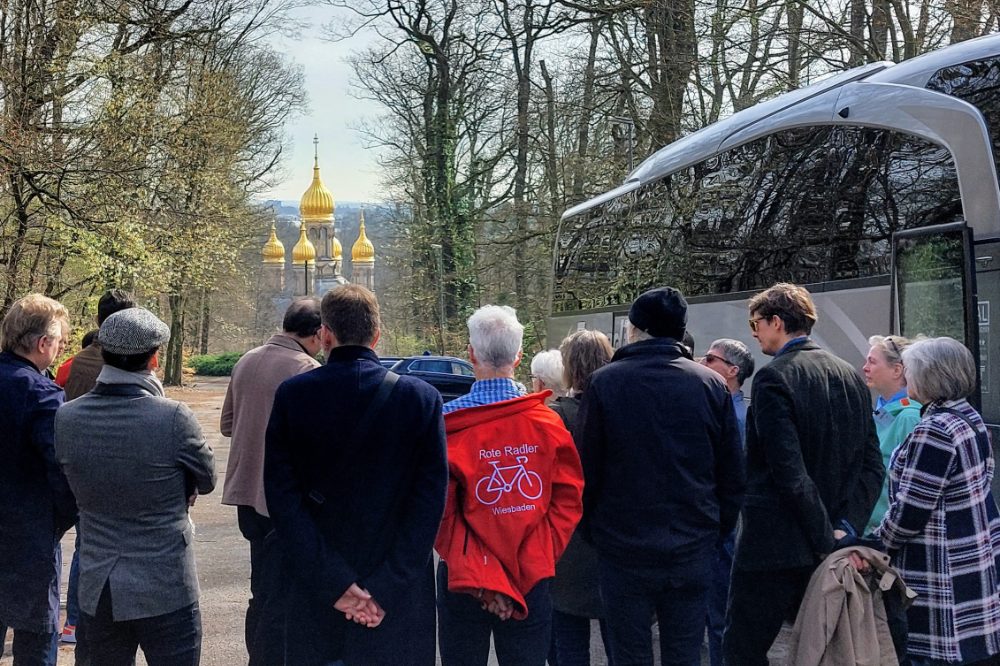 Eine Menschengruppe blickt an kahlen Laubbäumen vorbei auf goldene Kirchkuppeln. Neben den Menschen wartet ein Bus.