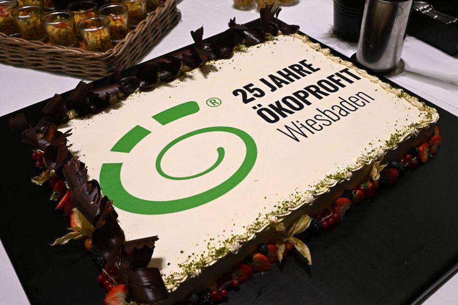 Правоъгълна торта с бяла глазура, зелено лого на Ökoprofit и текст "25 години Ökoprofit Wiesbaden", украсена с парченца шоколад и горски плодове.