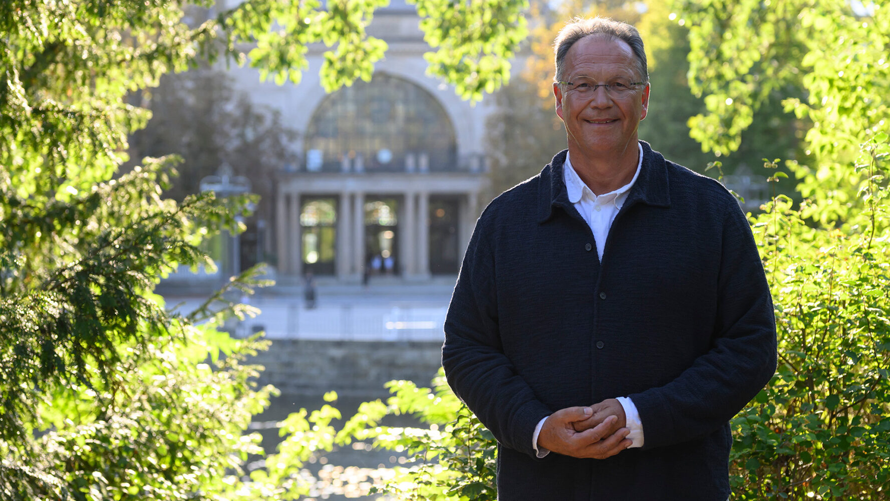 Man in front of a historic building with lots of greenery and water.