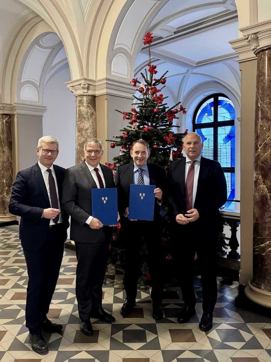 Auf dem Foto sind vier stehende Männer zu sehen, zwei halten eine blaue Mappe mit Lilien in der Hand. Im Hintergrund ist ein geschmückter Weihnachtsbaum zu sehen.