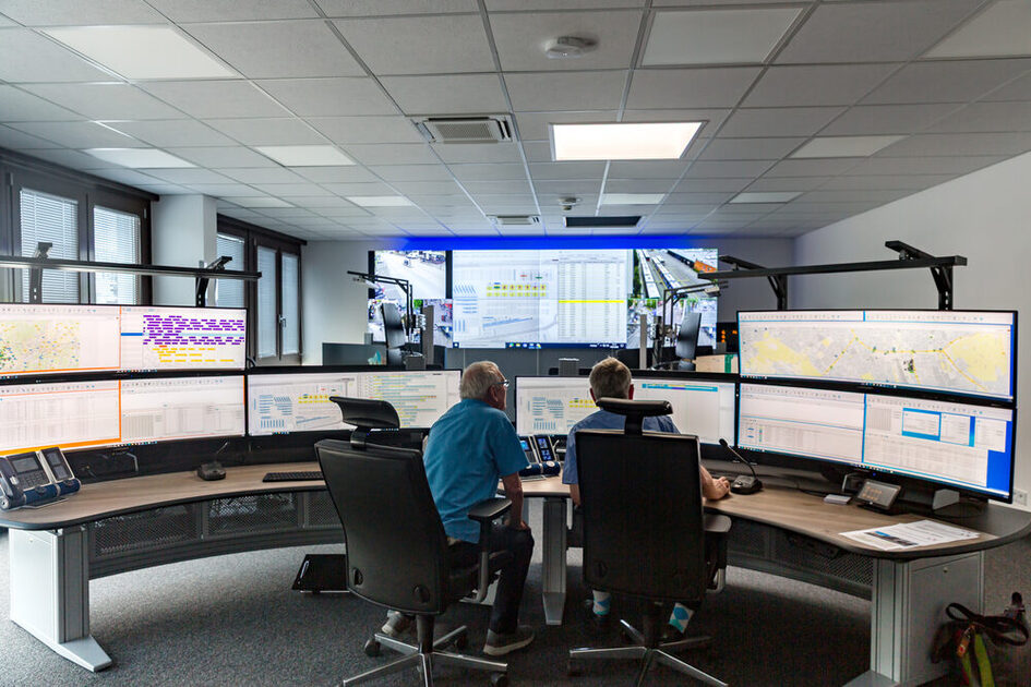 Es ist die Leitstelle mit vielen Bildschirmen und zwei Mitarbeitern, die am Schreibtisch sitzen, zu sehen.