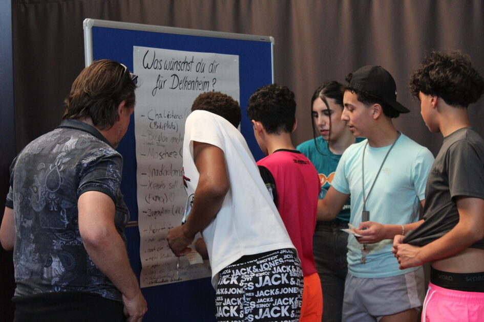 Jugendliche an einer Pinnwand mit einem Plakat, auf dem "Was wünschst du dir für Delkenheim?" steht.