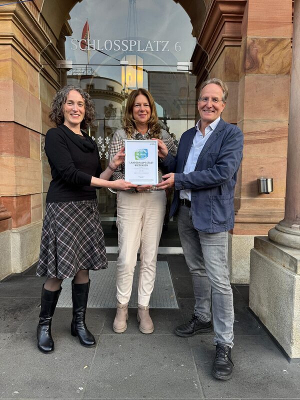 Es sind zwei Frauen und ein Mann vor einer Glastür zu sehen, die eine Urkunde in die Kamera halten.