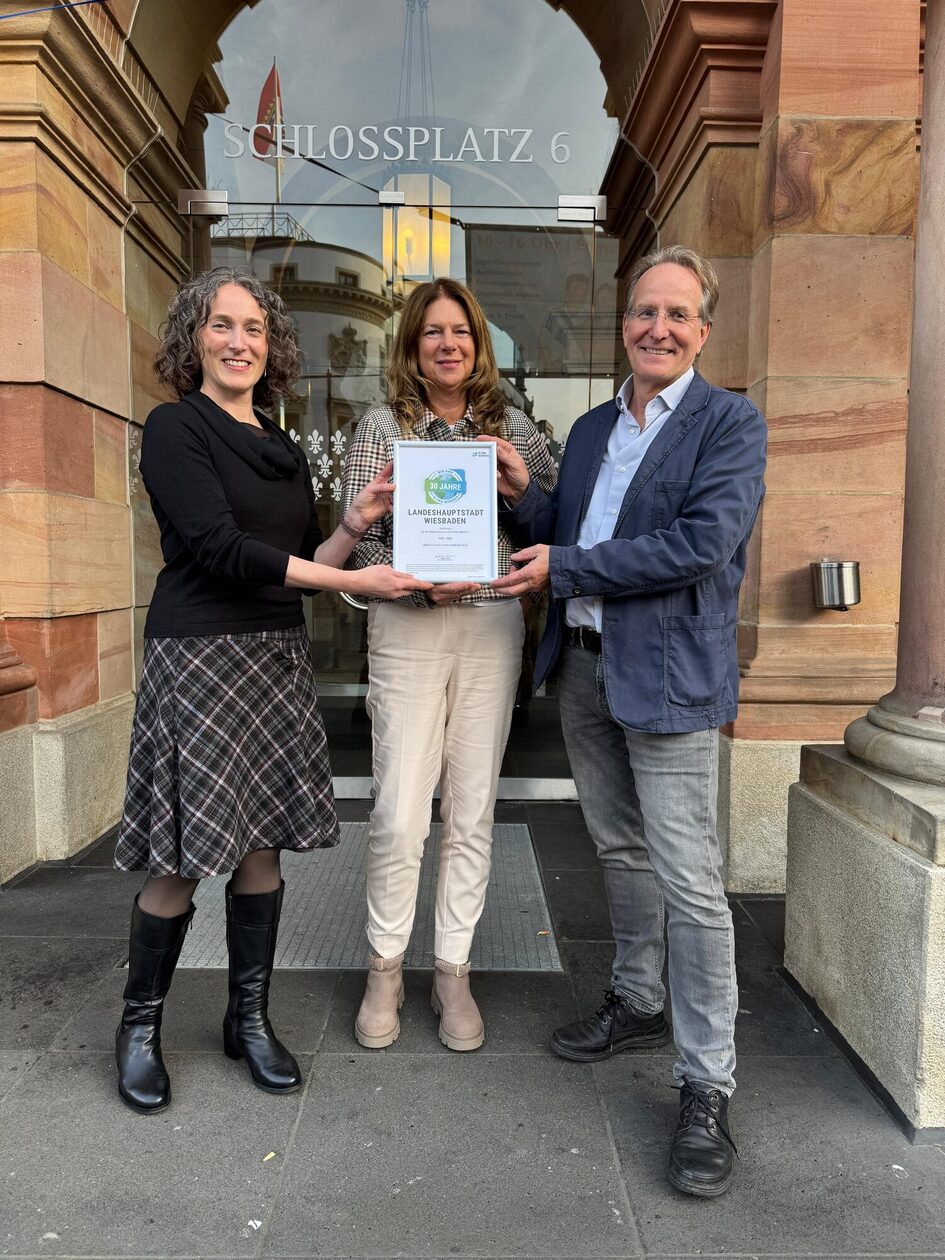 Es sind zwei Frauen und ein Mann vor einer Glastür zu sehen, die eine Urkunde in die Kamera halten.