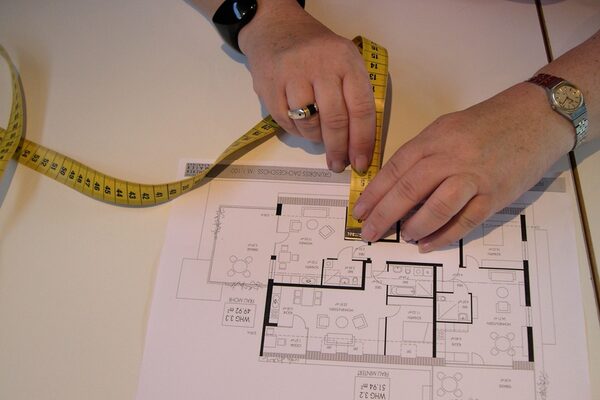 Auf dem Foto ist der Grundriss-Plan einer Wohnung zu sehen und zwei Hände, die mit einem Maßband Abstände messen.