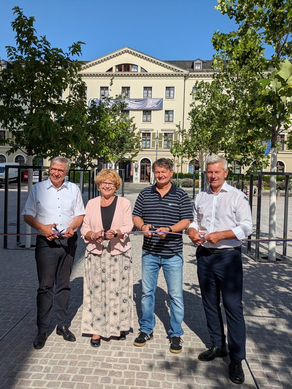 Auf dem Foto sind drei Herren und eine Dame auf dem Platz vor der Marktkirche zu sehen, die ein Band durchschneiden. Im Hintergrund sind Bäume und der Landtag zu sehen.