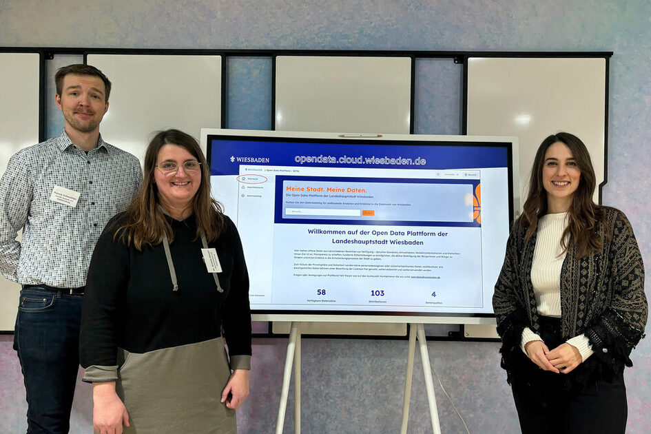 Auf dem Foto sind ein Mann und zwei Frauen vor einem digitalen Board zu sehen, das die Open-Data-Plattform der Landeshauptstadt Wiesbaden zeigt.