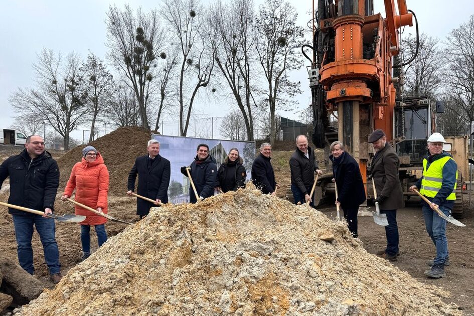 Auf dem Foto ist ein Erdhaufen zu sehen, im Hintergrund stehen zehn Personen mit Schaufeln.
