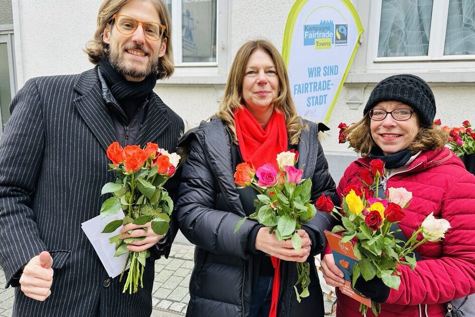 Auf dem Foto sind ein Mann und zwei Frauen in Winterkleidung auf einer Straße zu sehen, die jeweils Rosen in der Hand halten. Im Hintergrund ist eine Beachflag mit der Aufschrift "Wir sind Fairtrade-Stadt" zu sehen.