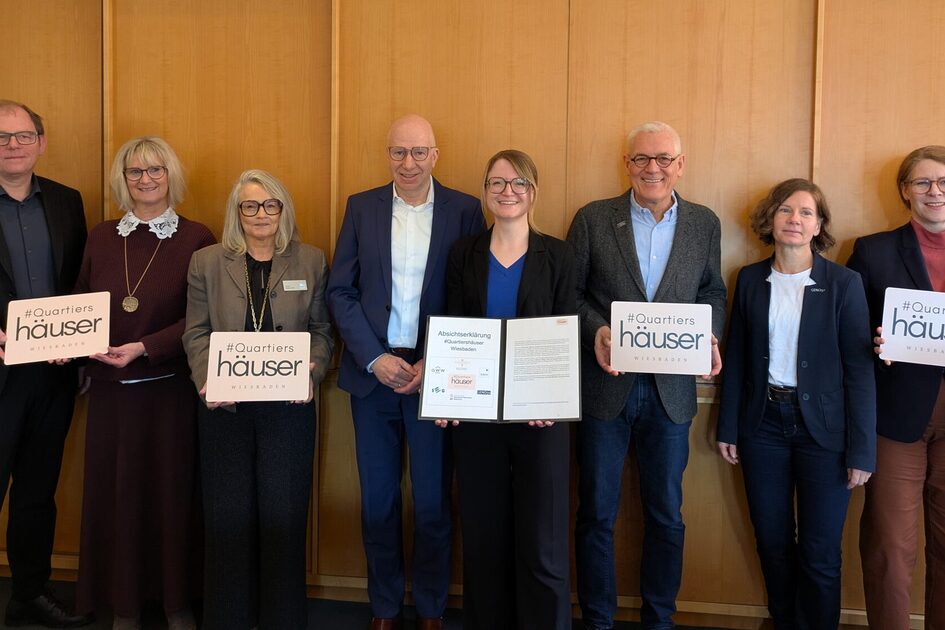 Auf dem Foto sind sieben Vertreterinnen und Vertreter der Wohnungsbaugesellschaften sowie Sozialdezernentin Dr. Becher zu sehen, die vor einer Holzwand stehen und Projektschilder sowie die unterzeichnete Absichtserklärung in den Händen halten.