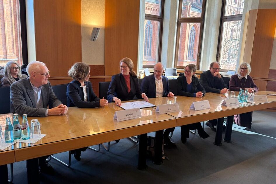 Auf dem Foto sitzen sieben Damen und Herren an einem länglichen Holztisch nebeneinander und unterzeichnen eine gemeinsame Erklärung.