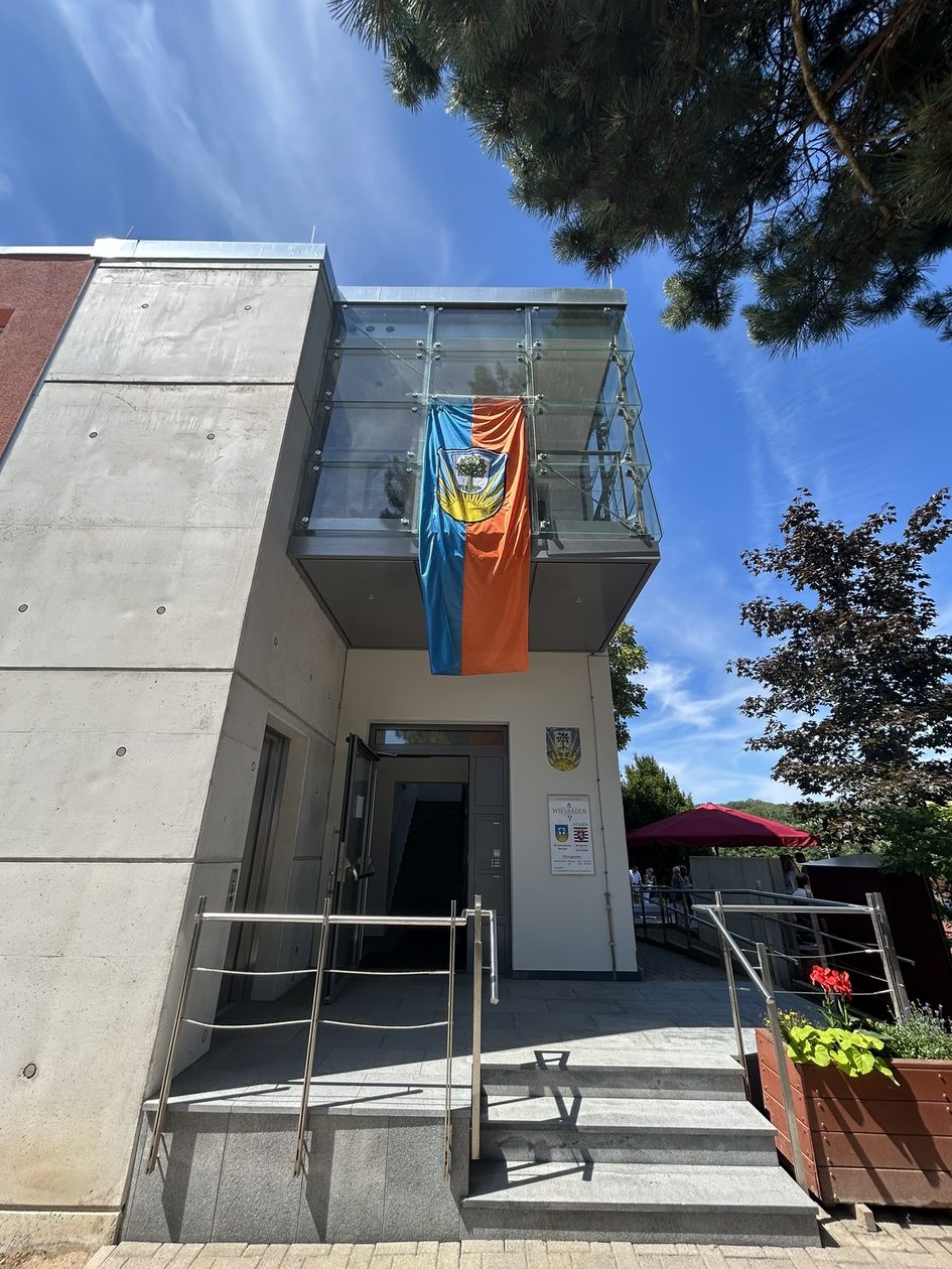 Auf dem Foto ist der Eingang der sanierten Ortsverwaltung Auringen zu sehen, an der eine blau-orangefarbige Flagge befestigt ist, die das Wappen von Auringen zeigt.