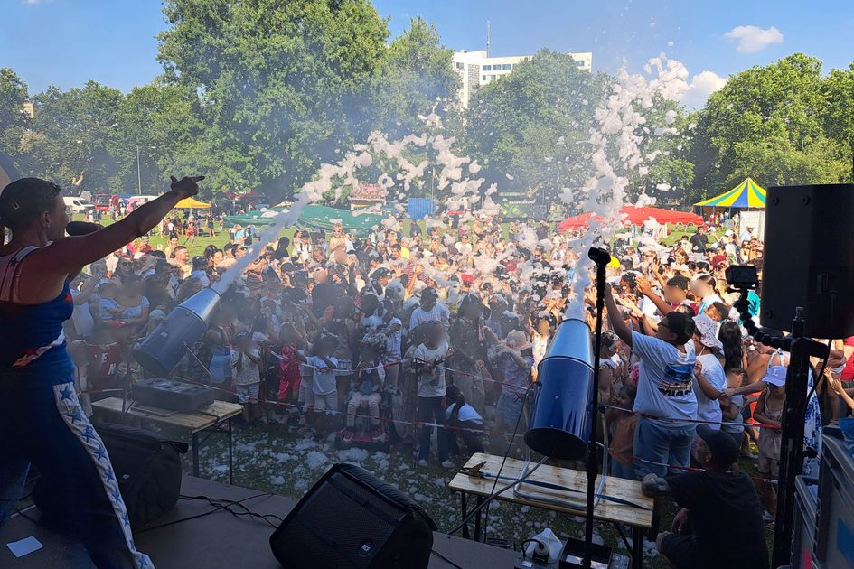 Das Foto wurde von der Bühne aufgenommen und zeigt eine Menschenmenge vor zwei Schaumwerfern, auf der Bühne ist von hinten eine singende Person zu sehen.