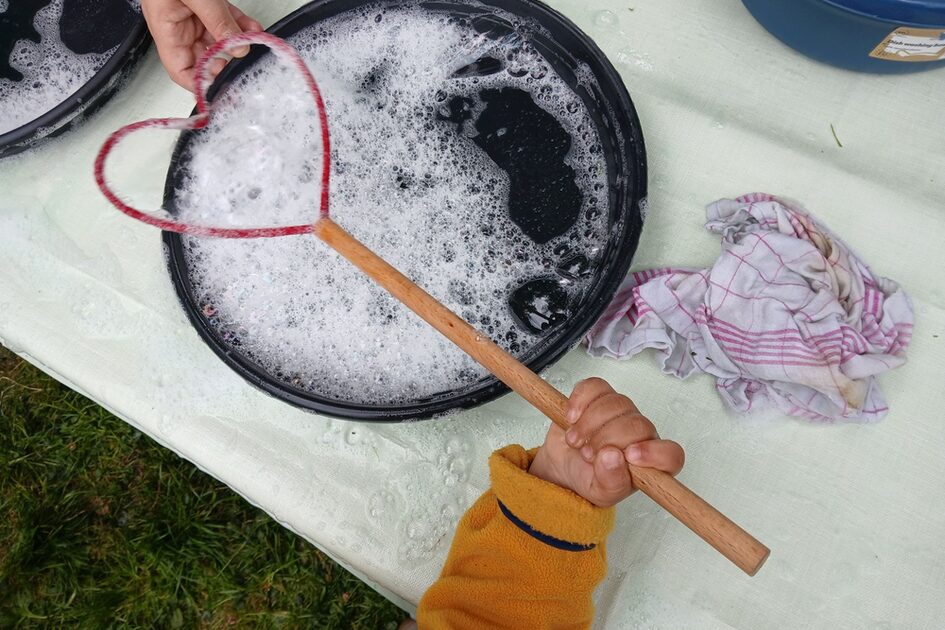 Auf dem Foto ist eine Kinderhand zu sehen, die einen herzförmigen Seifenblasenstab in der Hand hält, auf einem Tisch steht ein Eimer mit Seifenwasser.