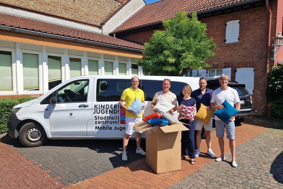 Auf dem Foto stehen vier Männer und eine Frau mit Kissen in der Hand zwischen einem Minivan der Jugendarbeit und einer Kiste mit weiteren Kissen.