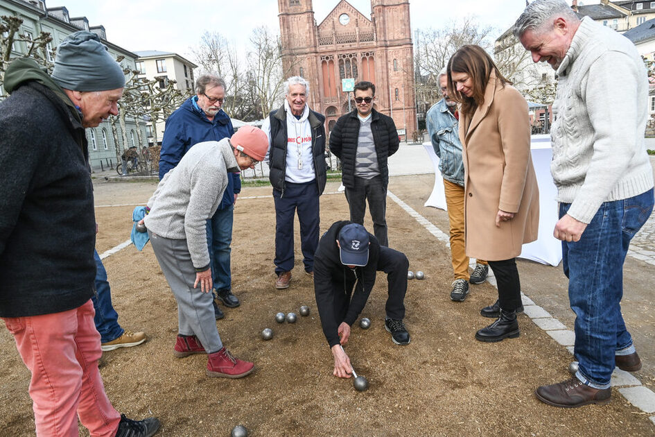 Auf dem Foto ist eine Gruppe Erwachsener zu sehen, die auf Boule spielen. Eine Person misst auf der Boulebahn den Abstand zwischen Kugeln.