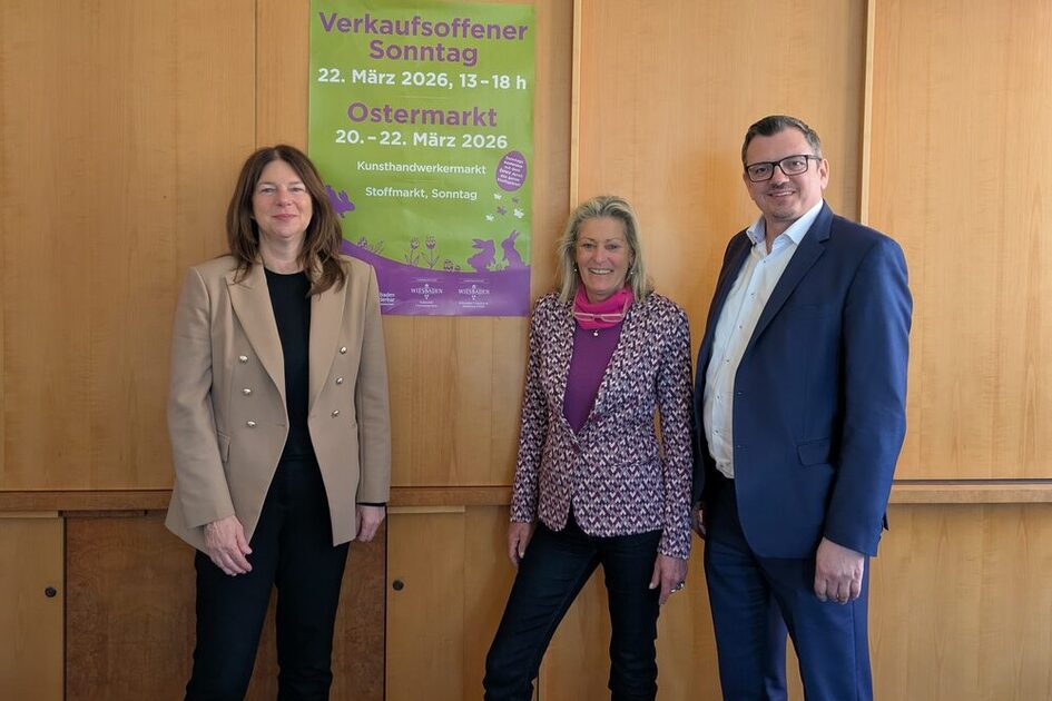 Auf dem Foto stehen zwei Damen und ein Herr vor einer Holzwand, an der ein grün-lilafarbenes Plakat zum Ostermarkt befestigt ist.