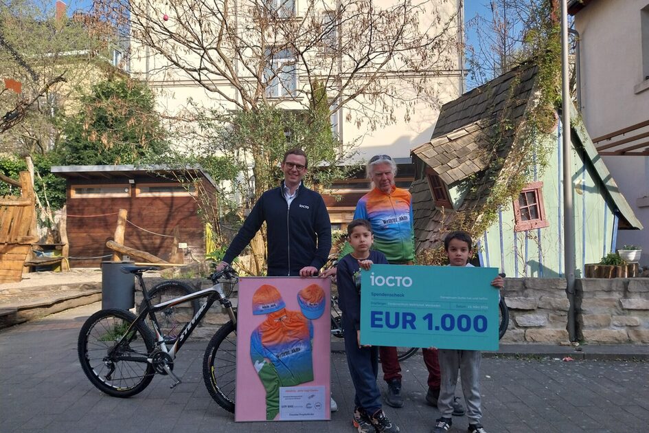 Das Foto zeigt zwei Männer und zwei Jungen, die mit einem Fahrrad und einem großen Spendenscheck im Innenhof des Kinderzentrums Wellritzhof stehen.