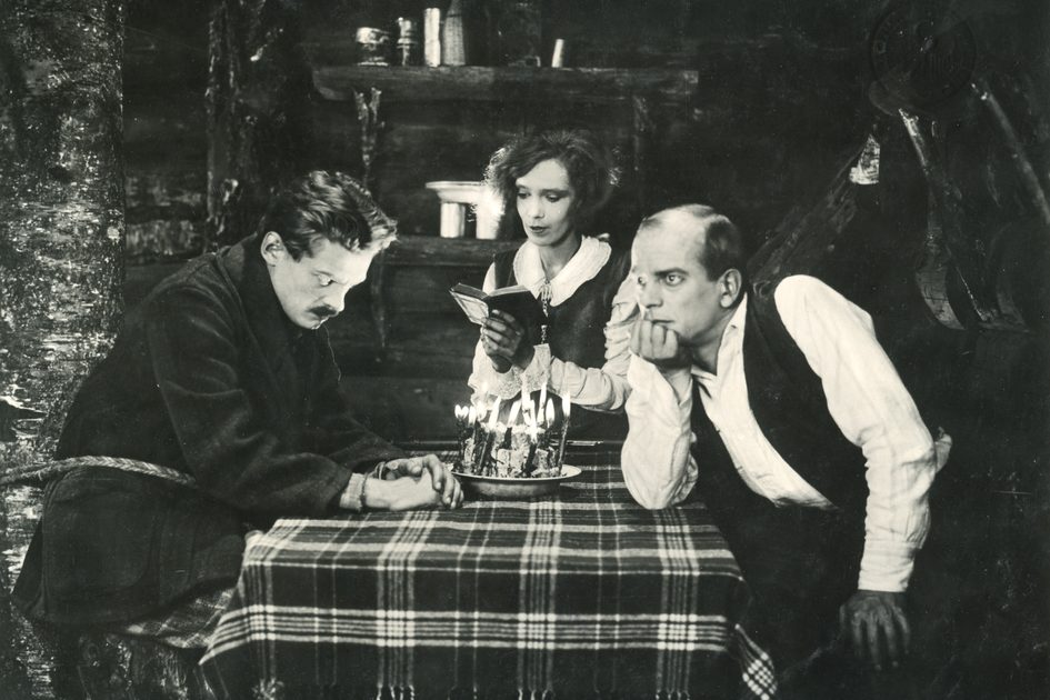 Stummfilmklassiker "Nach dem Gesetz": Es sind drei Personen in schwarz-weiß an einem Tisch zu sehen, auf dem Kerzen stehen.