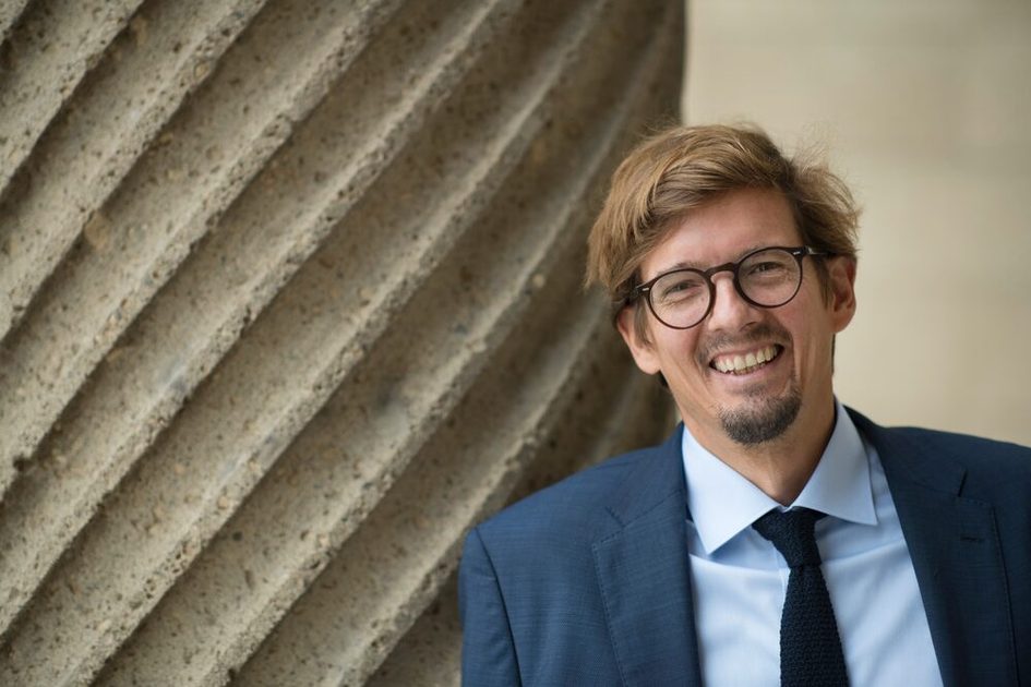 Bild von Camillo Huber-Braun in dunkelblauem Sakko, hellem Hemd und dunkler Krawatte, gut gelaunt lachend, vor einer steinernen Säule. Herr Huber-Braun trägt eine schwarze, runde Brille und über der Oberlippe sowie am Kinn einen kurzen Bart. Er hat hellbraune, kurze Haare.