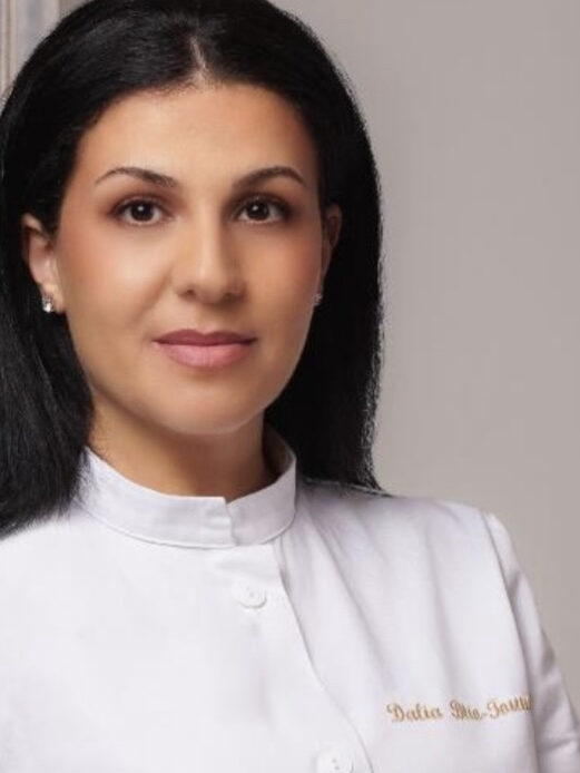 Kobieta z ciemnymi długimi włosami i białym topem uśmiecha się do kamery