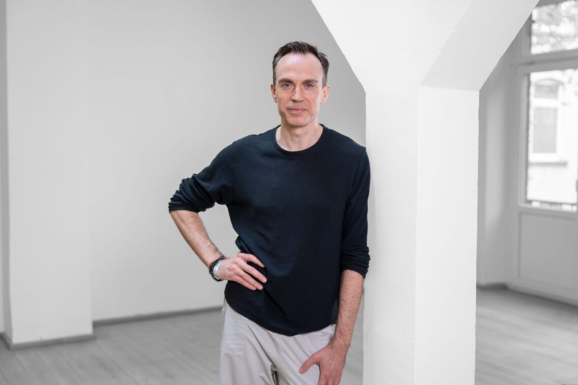 Мужчина с темной макушкой стоит в пустой комнате с белыми стенами и серым полом, прислонившись к белой колонне