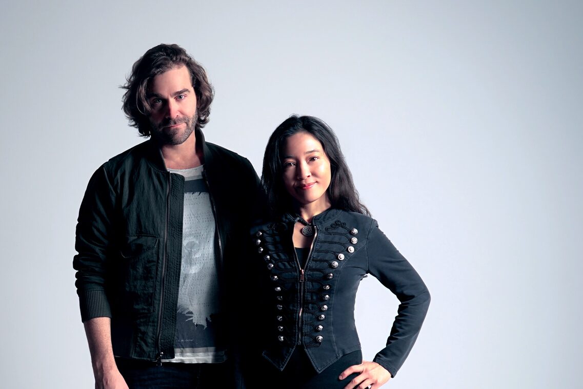 a man with long hair in a dark open jacket with a gray T-shirt underneath and a woman with a dark jacket with many buttons on both sides and long dark hair pose and look into the camera