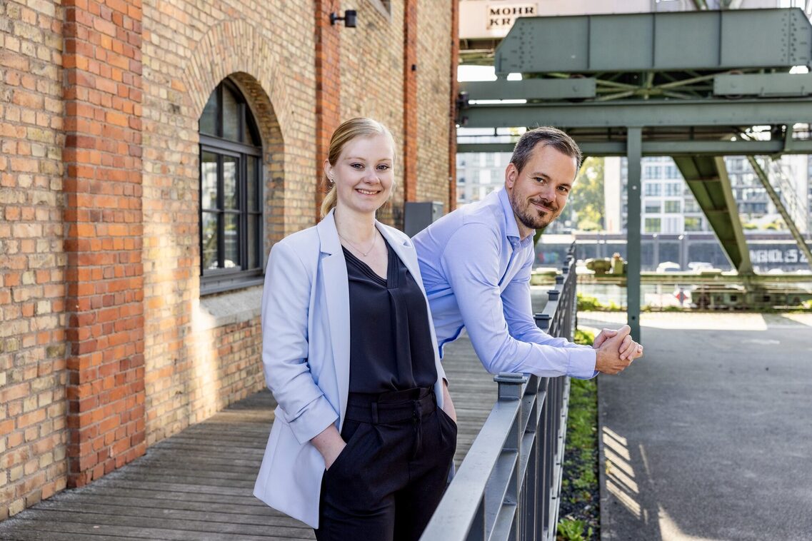 eine Dame und ein Herr stehen in die Kamera lächelnd vor einem Backsteingebäude auf einer Terrasse an dessen Geländer. Beide tragen hellblaue Oberteile, sie steht mit den Händen in den Taschen locker da, er lehnt mit beiden Armen vornüber hinter ihr auf dem Geländer.