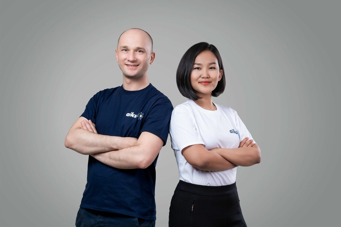 Ein Mann mit Glatze, dunkelblauem T-Shirt mit Aufschrift "aiku" auf der Brust und eine Frau mit halblangem dunklem Haar und weißem T-Shirt mit gleicher Aufschrift stehen nebeneinander und posieren mit verschränkten Armen in die Kamera lächeln.