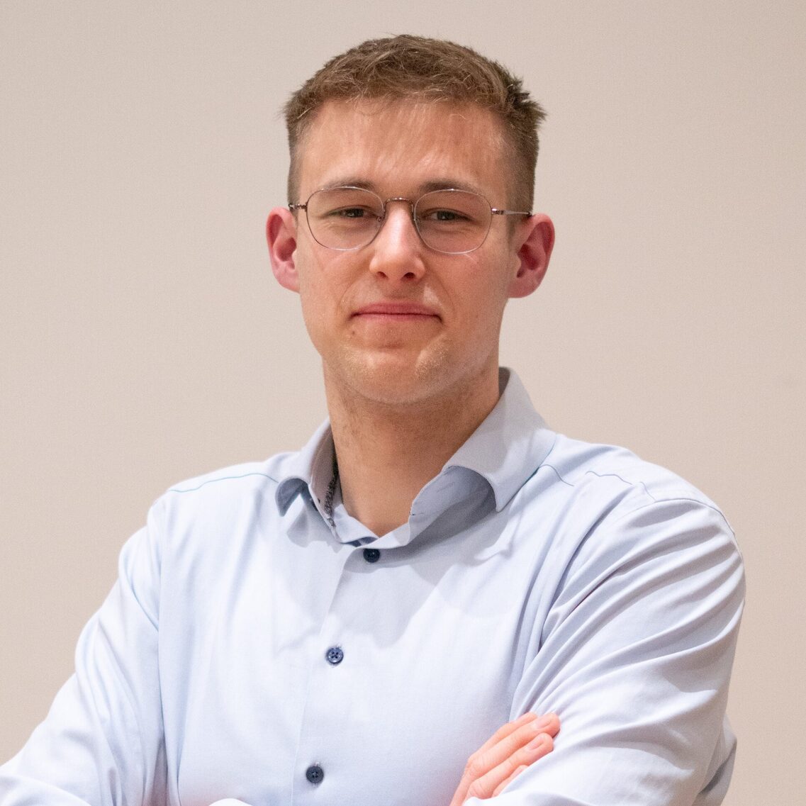 Άνδρας με γυαλιά και γαλάζιο πουκάμισο χαμογελάει στην κάμερα με σταυρωμένα τα χέρια του