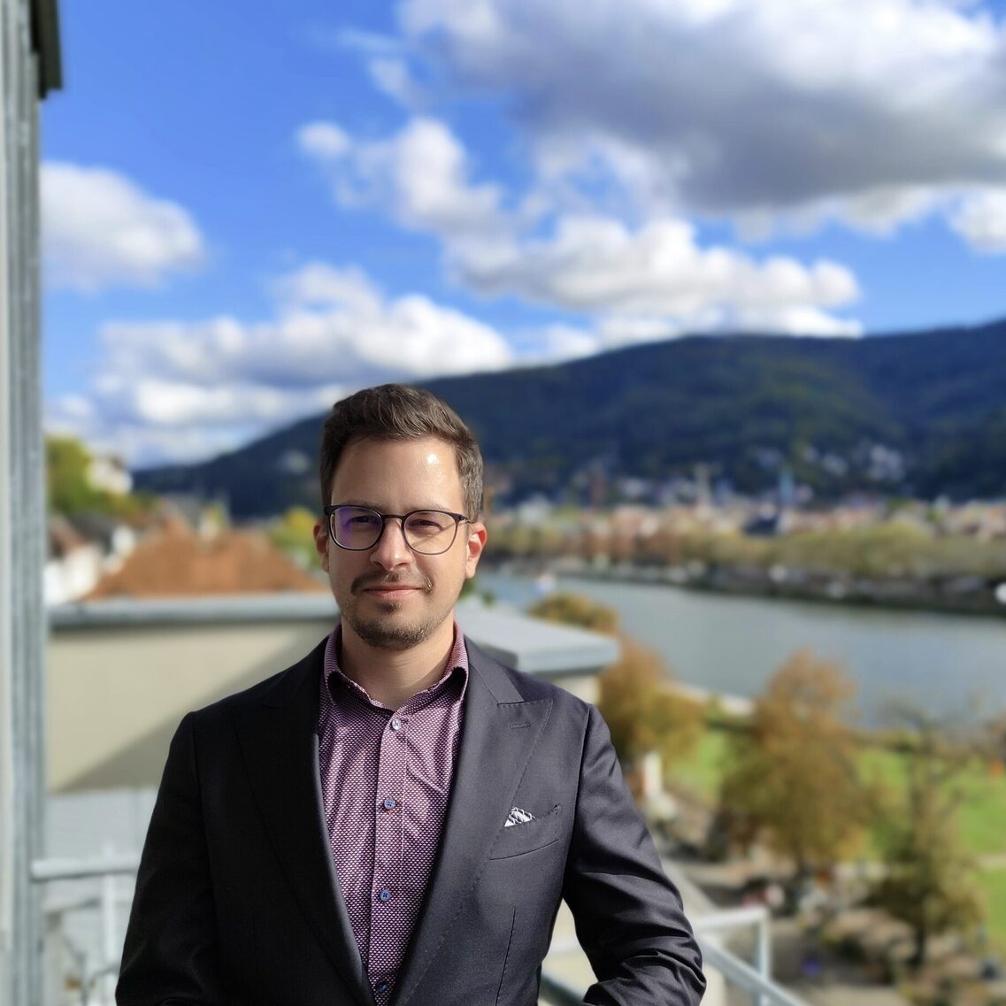 Herr in Anzug und Brille steht auf Balkon vor idyllischer städtischer Szenerie hinter Flusspanorama