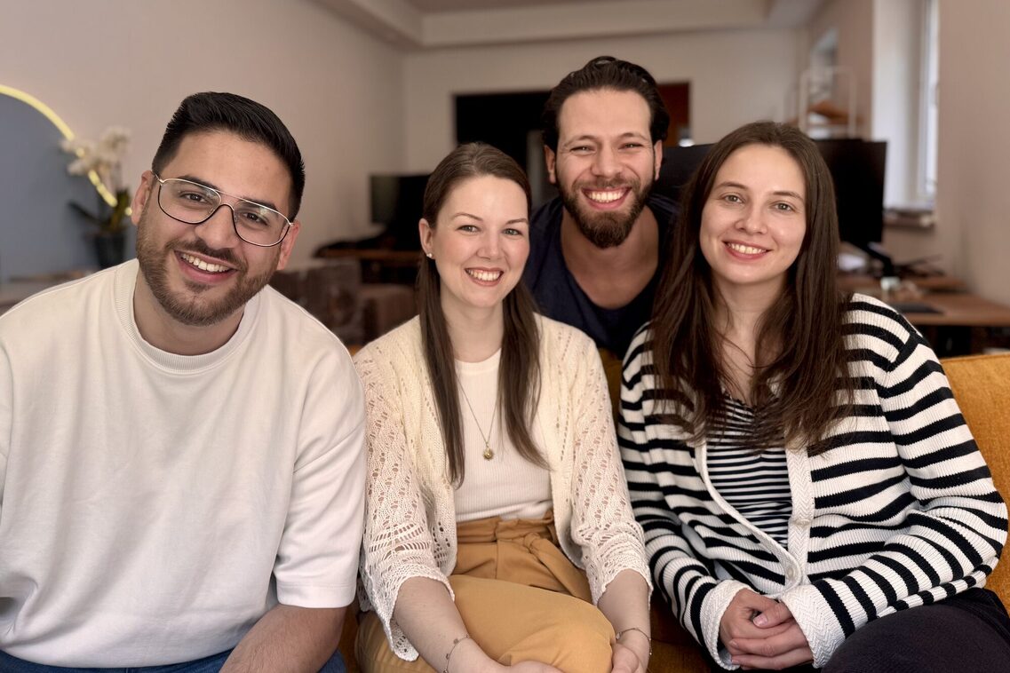 Vier junge Leute sitzen in einem Raum nebeneinander und lachen in die Kamera