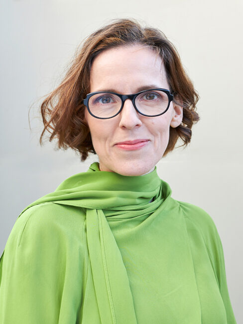 руда жінка з напівдовгим волоссям та окулярами посміхається в камеру у світло-зеленому топі на сірому тлі