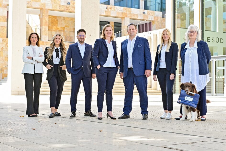 7 Damen und Herren im Businessdress und ein Hund mit Tasche im Maul posieren vor stilvollem Gebäude