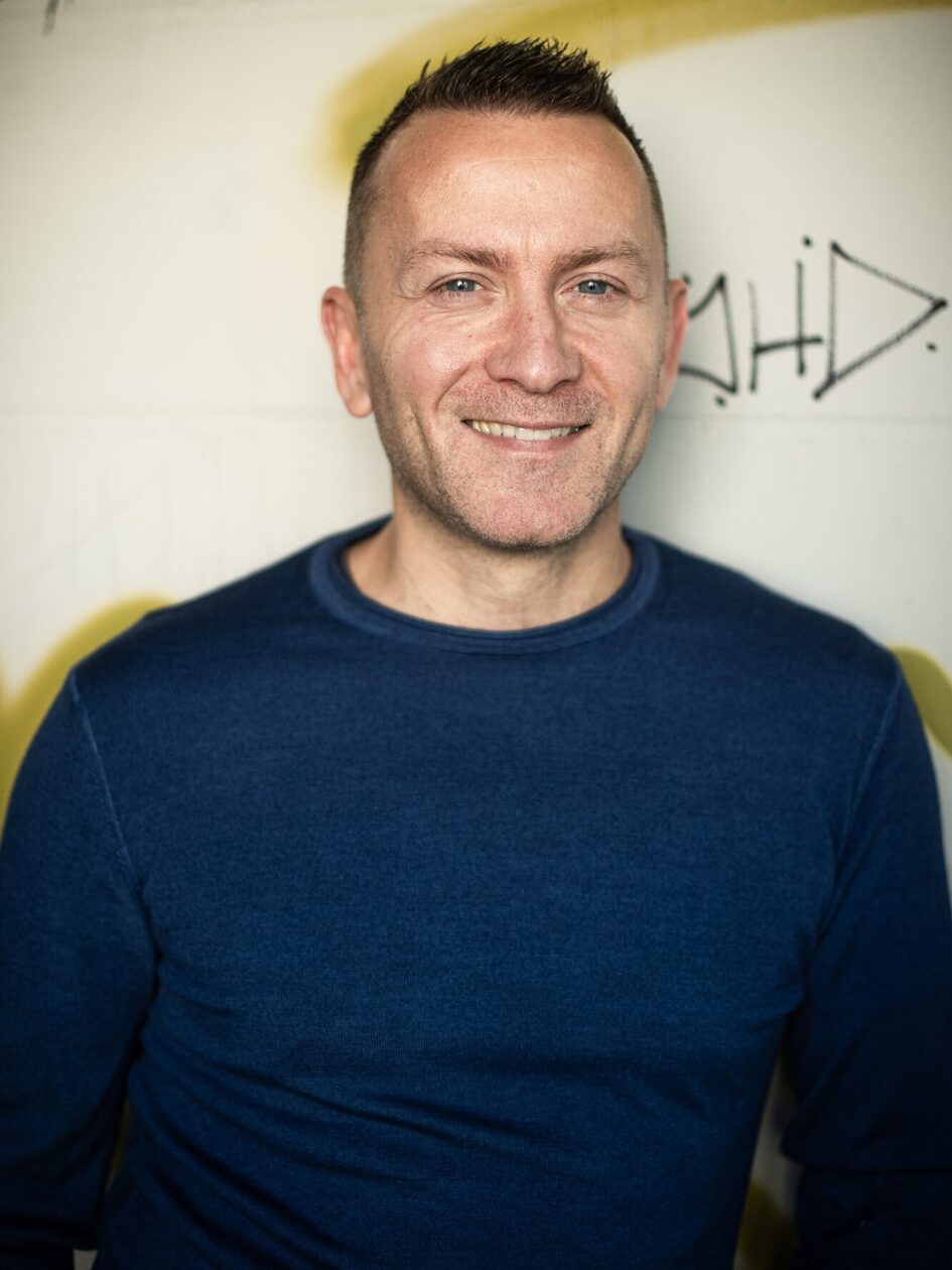 رجل يرتدي بلوزة زرقاء داكنة وشعر قصير أمام حائط مبلط عليه رسومات على الجدران يبتسم للكاميرا