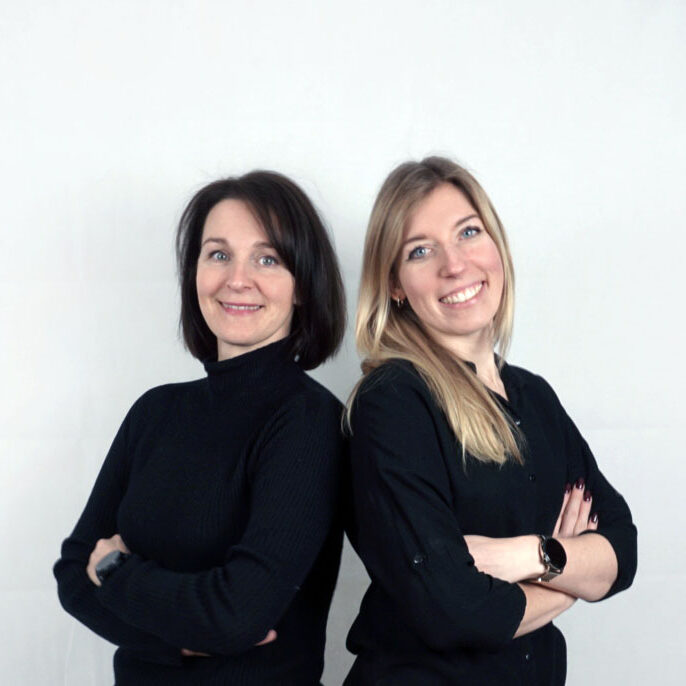 dwie kobiety stoją tyłem do kamery ze skrzyżowanymi ramionami, w czarnych bluzkach i uśmiechają się do siebie