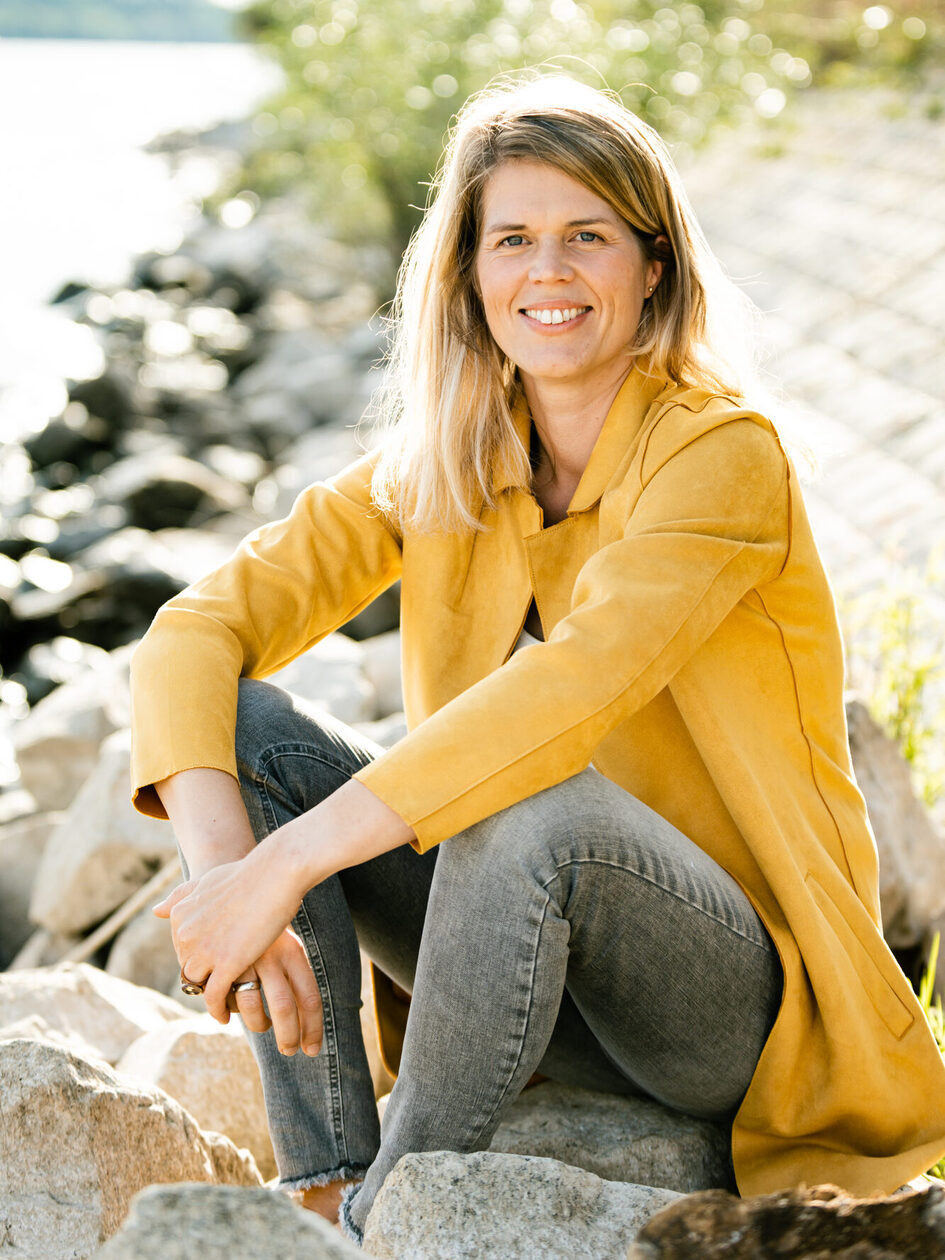 A woman in a yellow jacket sits on the stones of a riverbank and smiles into the camera