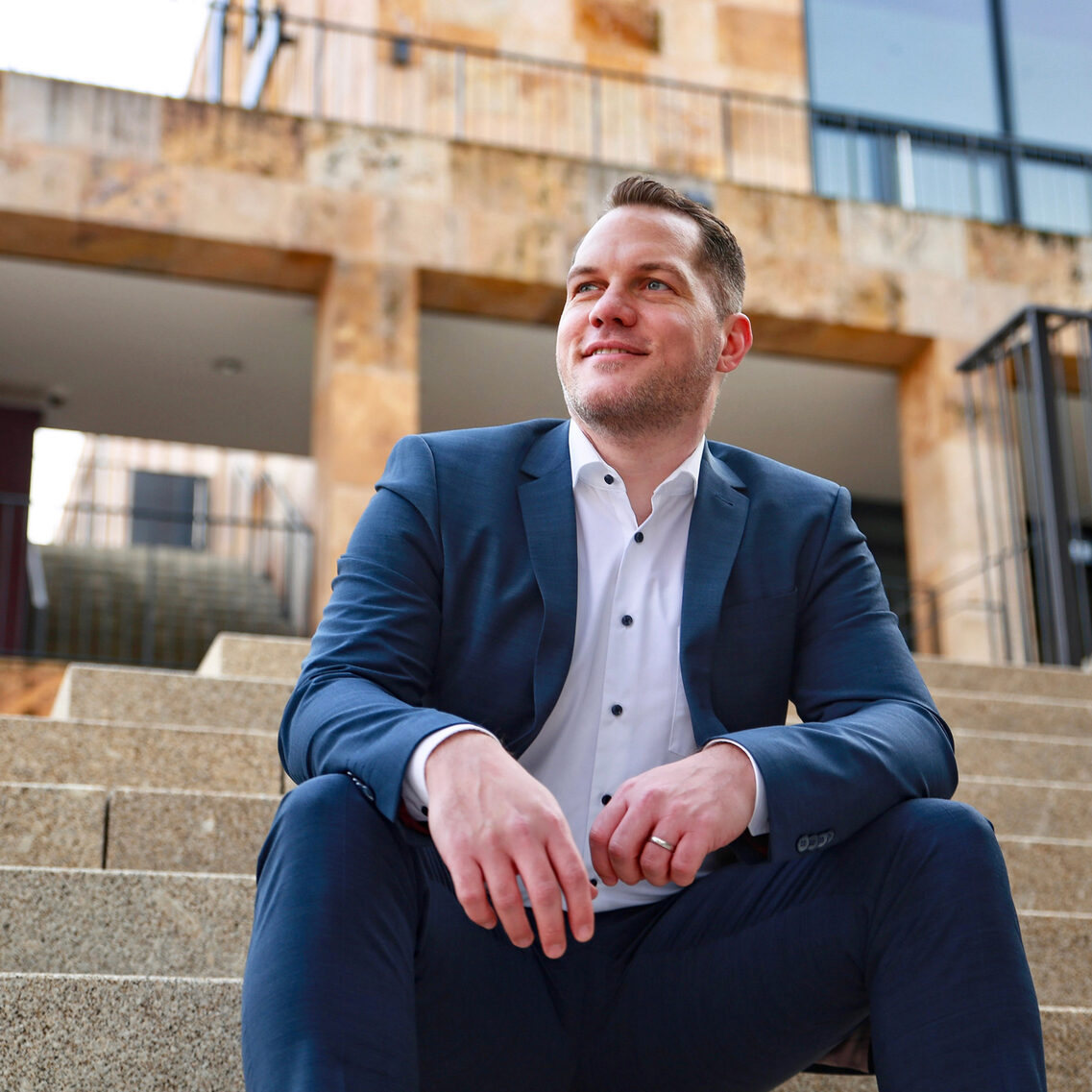 Mann in blauem Anzug sitzt auf Steintreppe und schaut lächelnd zur Seite