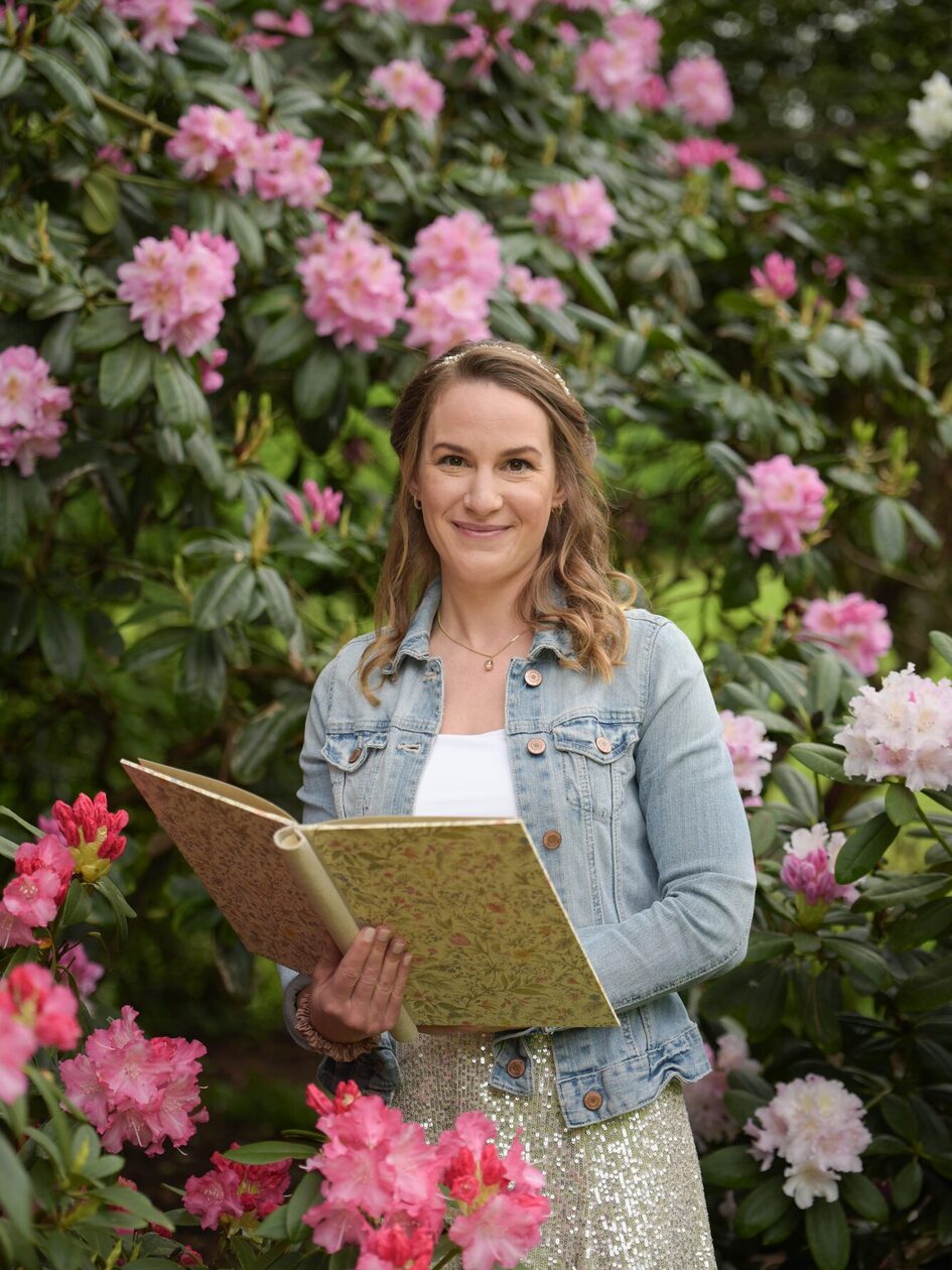 Frau hält aufgeschlagenes, verziertes, großes Buch in den Händen und schaut inmitten von Sträuchern mit weiß-pinken Blüten in die Kamera
