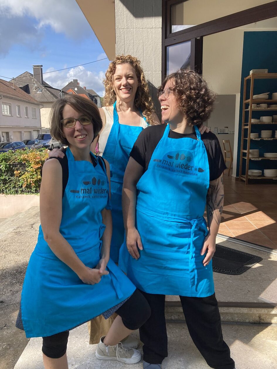 Tres mujeres de pie frente a una puerta de entrada abierta, con delantales azules y riéndose en parte a la cámara y en parte entre ellas.