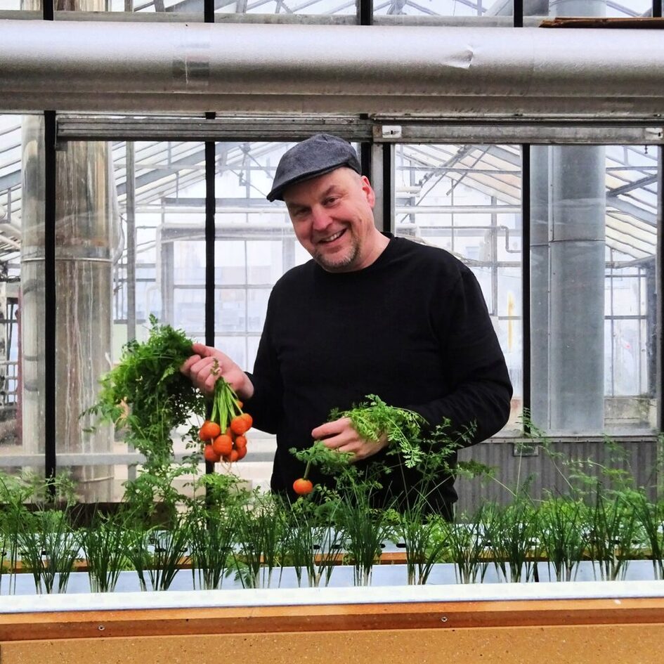 Mann mit Schiebermütze hält einen Bund radieschen in der Hand und steht hinter einem hydroponischem Beet